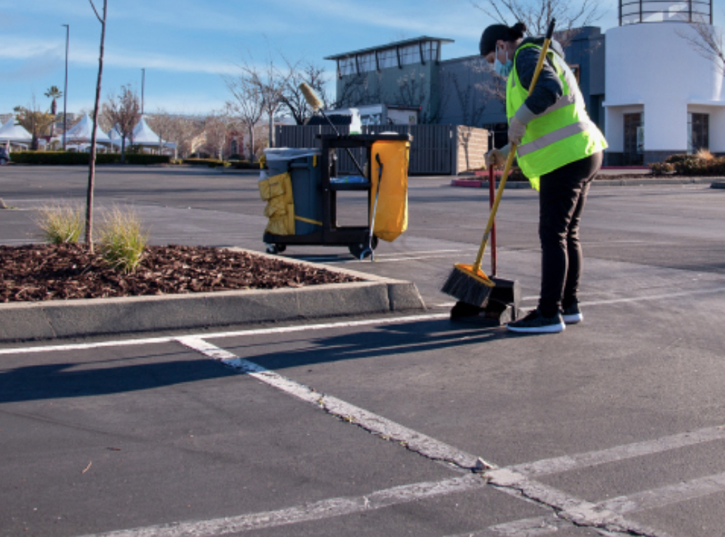 Worker in yellow safety vest sweeping a parking lot with a janitorial cart