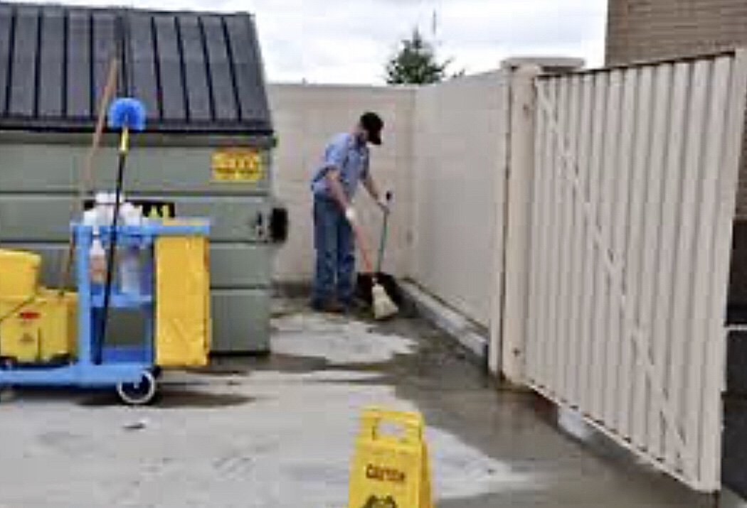 Worker cleaning behind a dumpster enclosure with janitorial cart